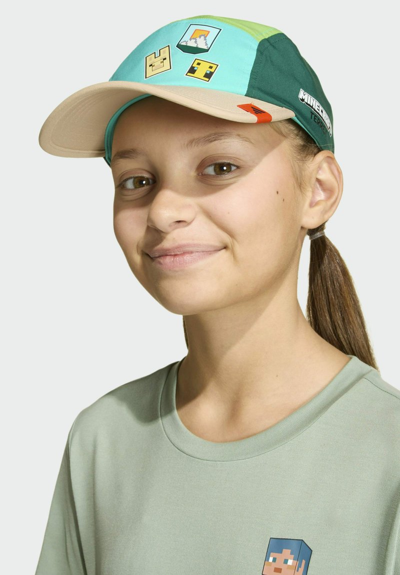 Młoda osoba w zielonej czapce z motywem Minecraft i pasującym jasnozielonym t-shircie, lekko się uśmiechająca z włosami związanymi z tyłu.