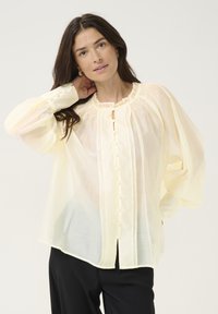 Blusa leggera di un giallo pallido, con maniche lunghe e a sbuffo, colletto arricciato e chiusura con bottoni sul davanti. Texture liscia, vestibilità comoda.