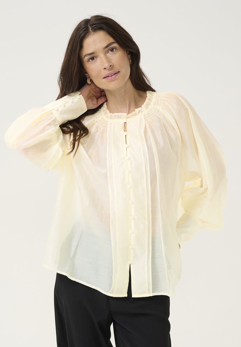 Blusa leggera di un giallo pallido, con maniche lunghe e a sbuffo, colletto arricciato e chiusura con bottoni sul davanti. Texture liscia, vestibilità comoda.