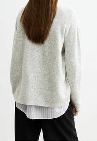 Dlouhé hnědé vlasy přes světle šedý svetr, který je vrstvený na bílé pruhované košili, doplněné �černými kalhotami, viděné zezadu.