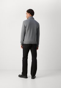 Grau gestrickter Pullover mit hohem Kragen, gerippten Bündchen und Saum, kombiniert mit schwarzen Jeans und schwarzen Schuhen, von hinten betrachtet.