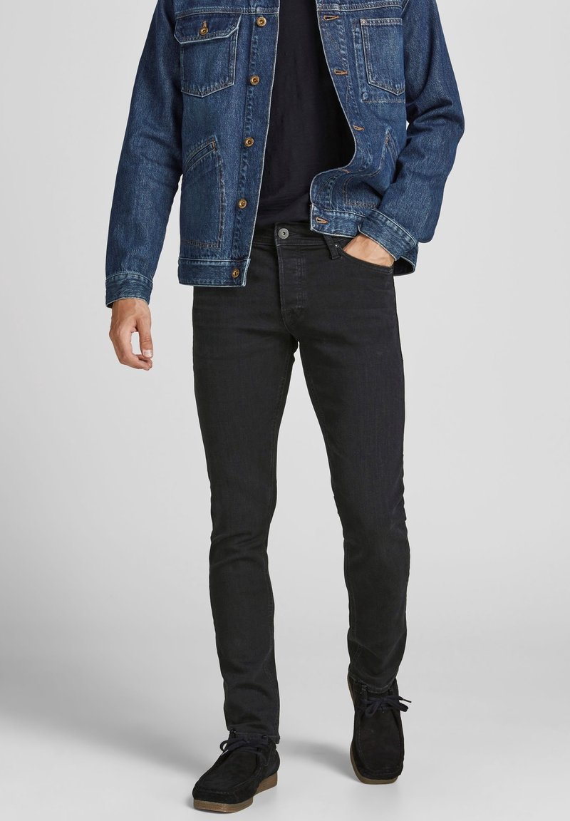 Homme portant une veste en jean bleue, un t-shirt noir, un jean slim noir et des chaussures noires à lacets sur un fond uni.