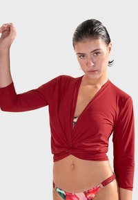 Crvena bluzica s omčom koja se veže sprijeda, s rukavima do 3/4 dužine i izvrnutim čvorom sprijeda, u kombinaciji s donjim dijelom bikinija s višebojnim cvjetnim uzorkom.