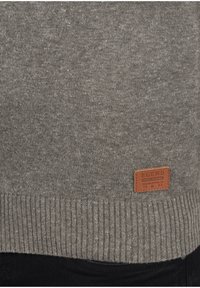 Maglione grigio a maglia con orlo a coste e un'etichetta in pelle marrone con la scritta "BLEND, Fondata nel 1993" in basso.