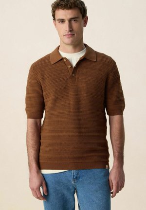 Jonge man die een bruin gestructureerd kortermig poloshirt draagt over een wit ondershirt en lichtblauwe spijkerbroek, staand tegen een effen achtergrond.