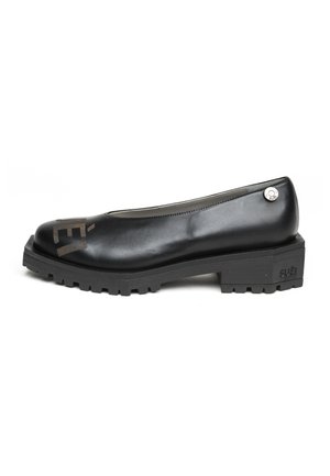 Scarpa piatta in pelle nera con suola spessa, scritta marrone sulla punta e piccolo logo metallico rotondo vicino al tallone su sfondo bianco.