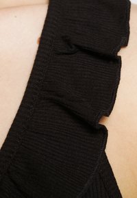 Svart ribbat tyg med volangkant. Texturen är slät med subtila åsar som framhäver en feminin design. Närbild.