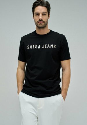 Hombre con cabello oscuro y barba, vistiendo una camiseta negra con el texto "SALSA JEANS" y pantalones blancos, de pie con las manos en los bolsillos frente a un fondo gris.