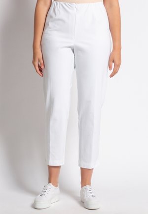Femme portant un pantalon blanc taille haute avec fermeture éclair à l'avant, longueur raccourcie, et des baskets blanches à plateforme lacées, debout devant un fond uni.