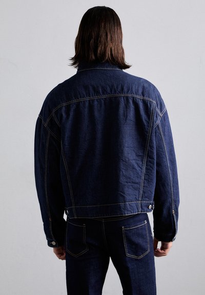 Denim jacket in dark blue with a relaxed fit. Features stitched details, a collared neckline, and buttoned cuffs. Matching dark denim pants are worn.