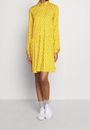 Femme portant une robe jaune à manches longues avec des motifs blancs en forme de losange, assortie à des chaussettes grises et des baskets blanches, debout devant un fond uni.