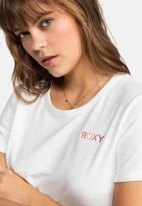 Camiseta blanca de manga corta fabricada con tejido ligero, que presenta un logotipo bordado de "ROXY" en tonos degradados en la zona del pecho.