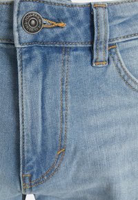 Ljust blå denimjeans med knäppning, orangea sömmar, bälteshällor och framfickor. Tyget har en något strukturerad yta.