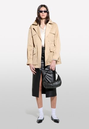 Veste en coton beige avec plusieurs poches, superposée à un chemisier en soie. Jupe en cuir noir avec une fente sur le côté. Les accessoires comprennent un sac noir et des lunettes de soleil.