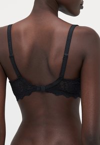 Black lace bra featuring a scalloped edge, adjustable straps, and a hook-and-eye closure at the back. Textured fabric with floral pattern.
