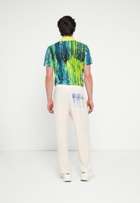 Többszínű póló absztrakt kék és zöld mintákkal, krémszínű laza nadrággal párosítva, hátul zsebfedős részlettel.