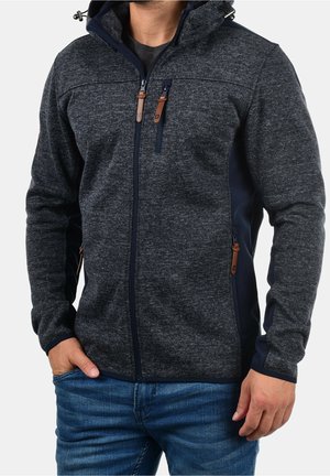 Mand iført mørkegrå hættetrøje med lynlås og tre lynlåslommer samt læderlynlåstræk, kombineret med blå jeans, hænderne afslappet langs siderne.