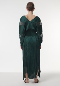 Vestido largo verde, con pliegues, espalda en V y mangas fluidas, hecho de una tela texturizada. Presenta una cintura ajustada y una abertura lateral.