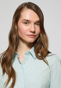 Camisa abotonada azul claro hecha de tejido suave, con cuello y un logotipo bordado en el lado izquierdo del pecho.