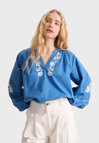 Blusa blu ricamata con scollo a V e maniche lunghe, caratterizzata da motivi floreali e dettagli bianchi. Abbinata a pantaloni di colore chiaro.