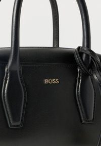 Μαύρη δερμάτινη τσάντα χειρός με δύο λαβές, λεπτομέρεια από δέρμα με κόμπο και χρυσό λογότυπο "BOSS" στην μπροστινή πλευρά.