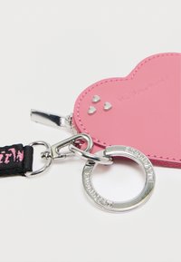 Ροζ θήκη σε σχήμα καρδιάς κατασκευασμένη από απαλό δέρμα με ασημένια υλικά. Διαθέτει φερμουάρ και μαύρο ιμάντα με ροζ γράμματα. Τρία μικρά διακοσμητικά σε σχήμα καρδιάς.