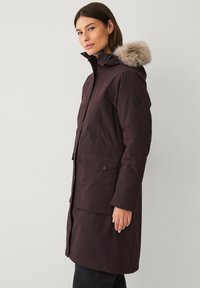 Bruine parka tot boven de enkels met een capuchon met bontafwerking, twee voorzakken, rits- en drukknoopsluitingen, en een zwart rond logo op de mouw.