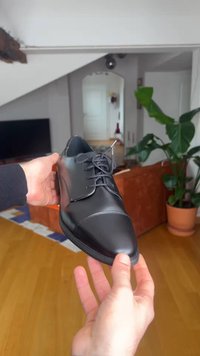 Mains tenant une chaussure habillée noire en cuir poli avec lacets dans un salon avec un sol en bois, un canapé, une plante et un miroir.