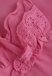 Rosa plagg med en strukturert blondevolang langs halsen, med glatt stoff i samme nyanse. Det overordnede designet er mykt og delikat.