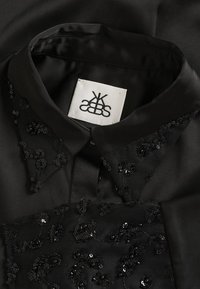 Crna bluzica od tkanine koja sadrži ovratnik ukrašen crnim šljokicama, teksturiranim detaljima i etiketom sa stiliziranim logotipom.