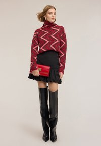 Červený a růžový vzorovaný rolák, černá sukýnka s volánky a vysoké černé boty s špičatou špičkou, držící červenou psaníčko s zlatým emblémem.