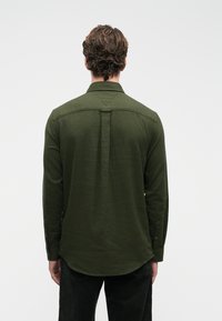 Σκοτεινό πράσινο πουκάμισο με μακριά μανίκια και λεπτομέρεια στο πίσω μέρος, καμπυλωτό στρίφωμα και κουμπωτές μανσέτες. Το ύφασμα φαίνεται λείο με ελαφριά υφή.