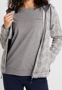 Veste grise à fermeture éclair avec un tissu à motifs sur les manches, sweat-shirt gris en dessous, et un design simple. Comprend des couches bicolores et des fermetures éclair.