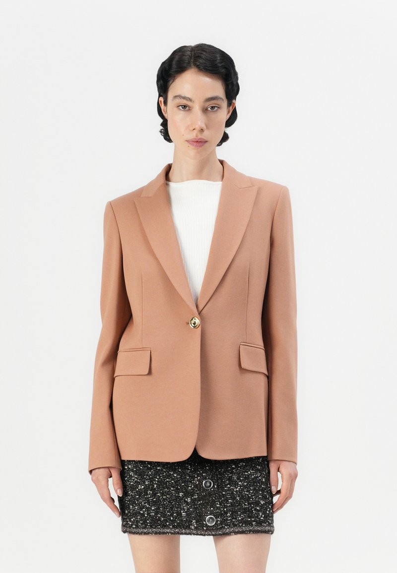 Brzoskwiniowa marynarka w dopasowanym fasonie, z klapami i dwoma przednimi kieszeniami, zapinana na złoty guzik. Noszona na białej bluzce i czarnej teksturowanej spódnicy.