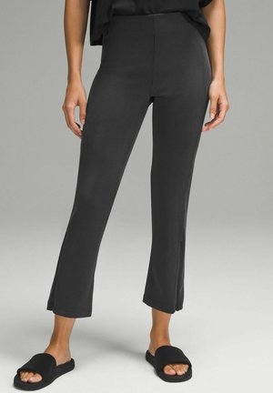 Femme portant un pantalon noir court côtelé et des chaussons noirs à bout ouvert, debout devant un fond gris uni.