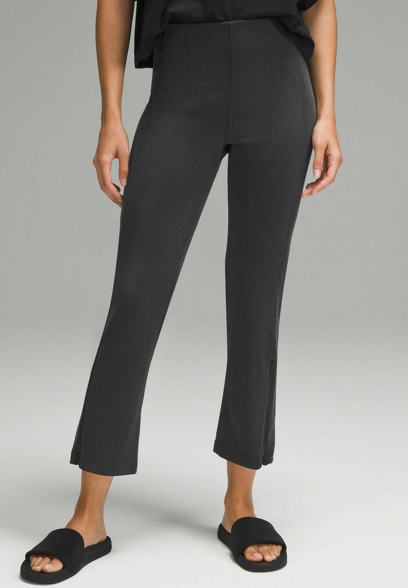 Femme portant un pantalon noir court côtelé et des chaussons noirs à bout ouvert, debout devant un fond gris uni.