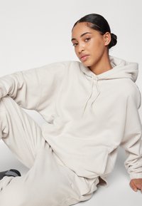 Felpa oversized crema con cappuccio regolabile, in morbido pile testurizzato e un logo sottile sul petto, abbinata a pantaloni jogging coordinati.