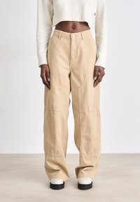 Pantalons larges beiges en tissu résistant, avec une taille haute, des coutures visibles et des poches avant. Les chaussures sont des bottes à semelles épaisses.