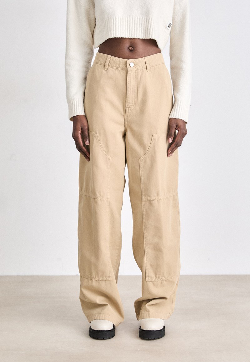 Pantalons larges beiges en tissu résistant, avec une taille haute, des coutures visibles et des poches avant. Les chaussures sont des bottes à semelles épaisses.