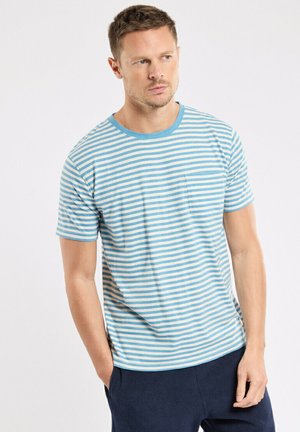 Homme aux cheveux courts portant un t-shirt rayé bleu clair et blanc et un pantalon foncé, debout les mains dans les poches, regardant sur le côté.