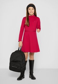 Piros, hosszú ujjú ruha, amelynek karcsúsított derékrésze és csillogó dísze van, fekete térdig érő csizmával és fekete háti táskával párosítva, fehér logóval.