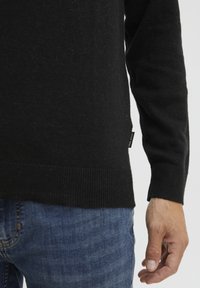 Fekete kötött pulóver bordás mandzsettákkal, sima textúrával és egy kis címkével az alján. Kék farmernadrággal viselve.