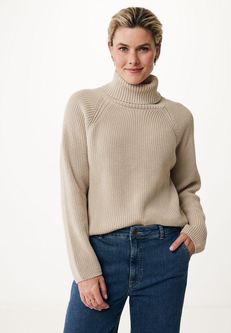 Beige knit sweater with a high collar, ribbed texture, and raglan sleeves, paired with dark blue denim jeans. Simple design, relaxed fit.