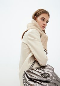 Mladá žena s dlouhými vlasy má na sobě béžový kabát a oversized šálu, drží kovovou kabelku s řetízkovým popruhem na bílém pozadí.