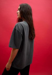 Przesadny szary t-shirt z krótkimi rękawami, okrągłym dekoltem i miękką fakturą, wyeksponowany na jednolitym czerwonym tle.