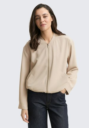 Veste bomber beige avec fermeture éclair sur le devant, deux poches latérales et manches longues. Texture lisse et coupe décontractée, associée à un pantalon en denim foncé.