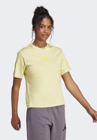 Žluté sportovní tričko s krátkým rukávem a kulatým výstřihem, s jemným trojúhelníkovým logem ve světlejším odstínu, v kombinaci se šedými kalhotami.