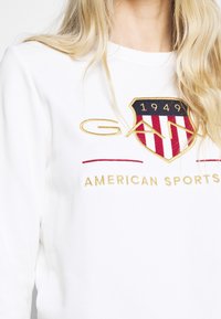 Bílá mikina s vyšívaným logem s červeno-modrým štítovým vzorem, zlatými akcenty a textem "AMERICAN SPORTS" pod ním.