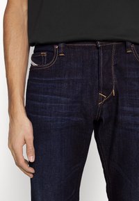 Jeans de mezclilla en azul oscuro con costuras naranjas, un diseño clásico de cinco bolsillos y una textura sutil. La cintura incluye un cierre de botón y cremallera.