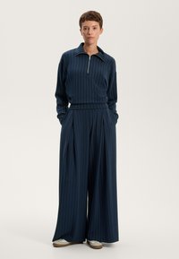 Temno modra trenerka z navpičnimi belimi črtami; zadrga pri ovratu in široke hlače; mehak material z sproščenim krojem.